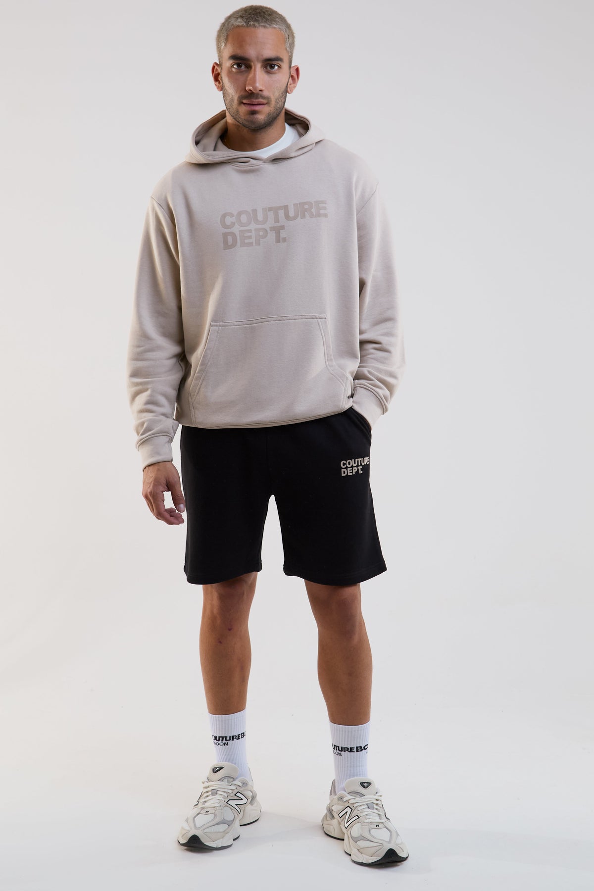 Man wearing a beige hoodie and black shorts with 'Couture Dept.' branding on a white background