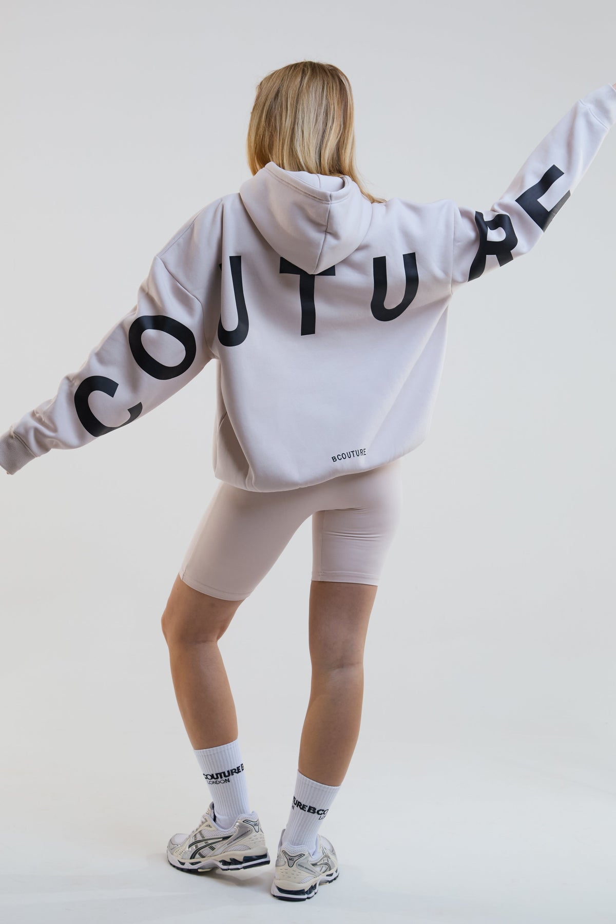 Person wearing a beige hoodie with 'COUTURE' text on the back, standing against a plain background.