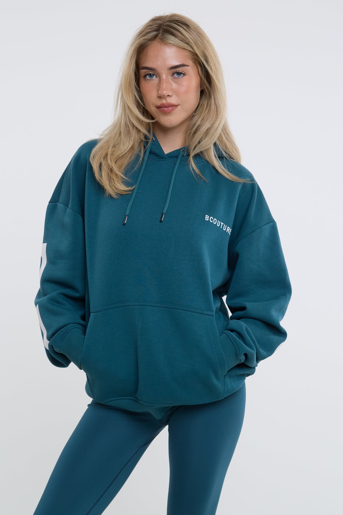 A blonde woman wearing an oversized teal blue-green hoodie with white text on the chest, paired with matching leggings, against a light grey studio background.