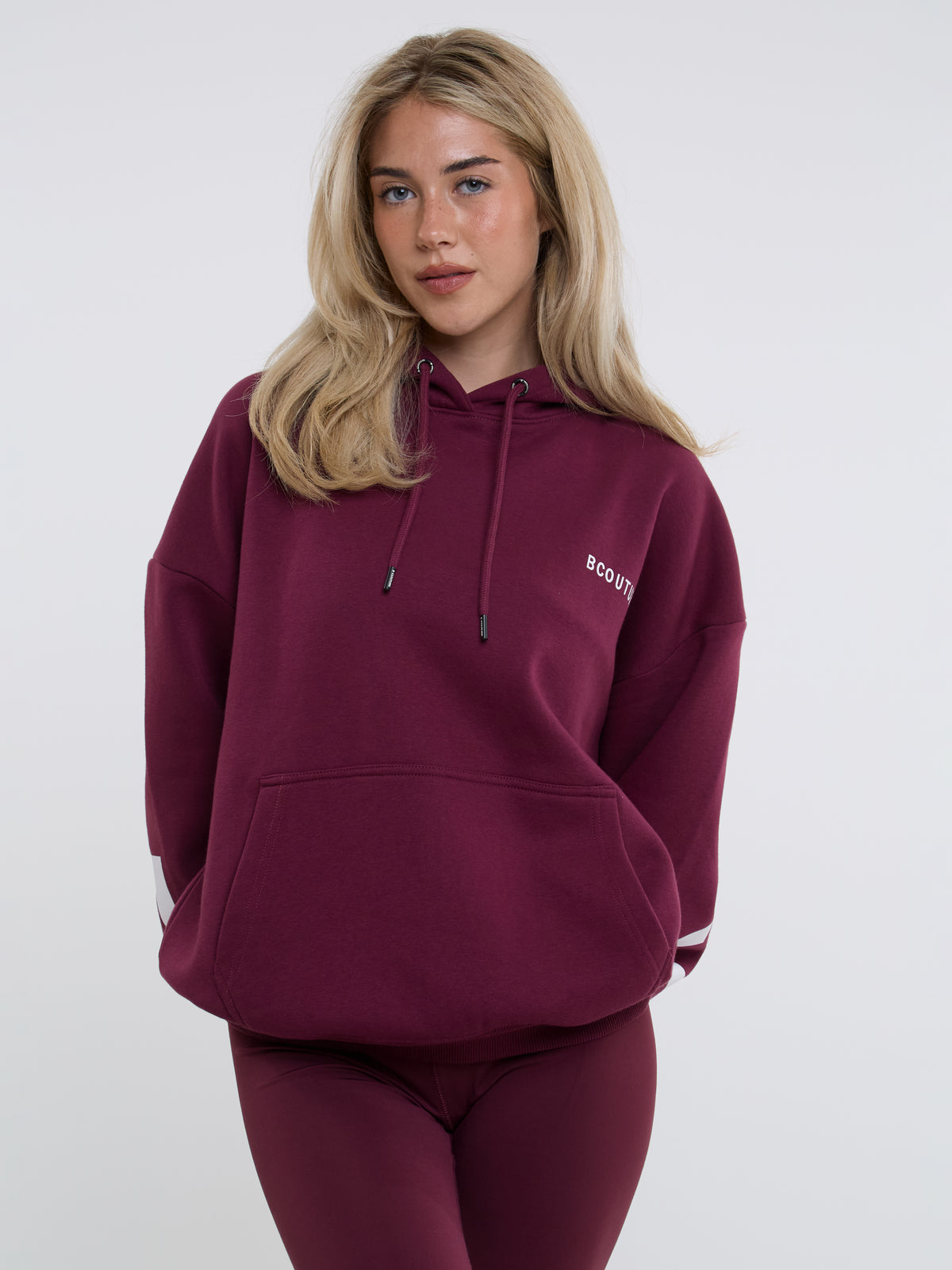 A woman wearing an oversized burgundy pullover hoodie with a drawstring hood and kangaroo pocket, paired with matching leggings, against a light gray studio background.