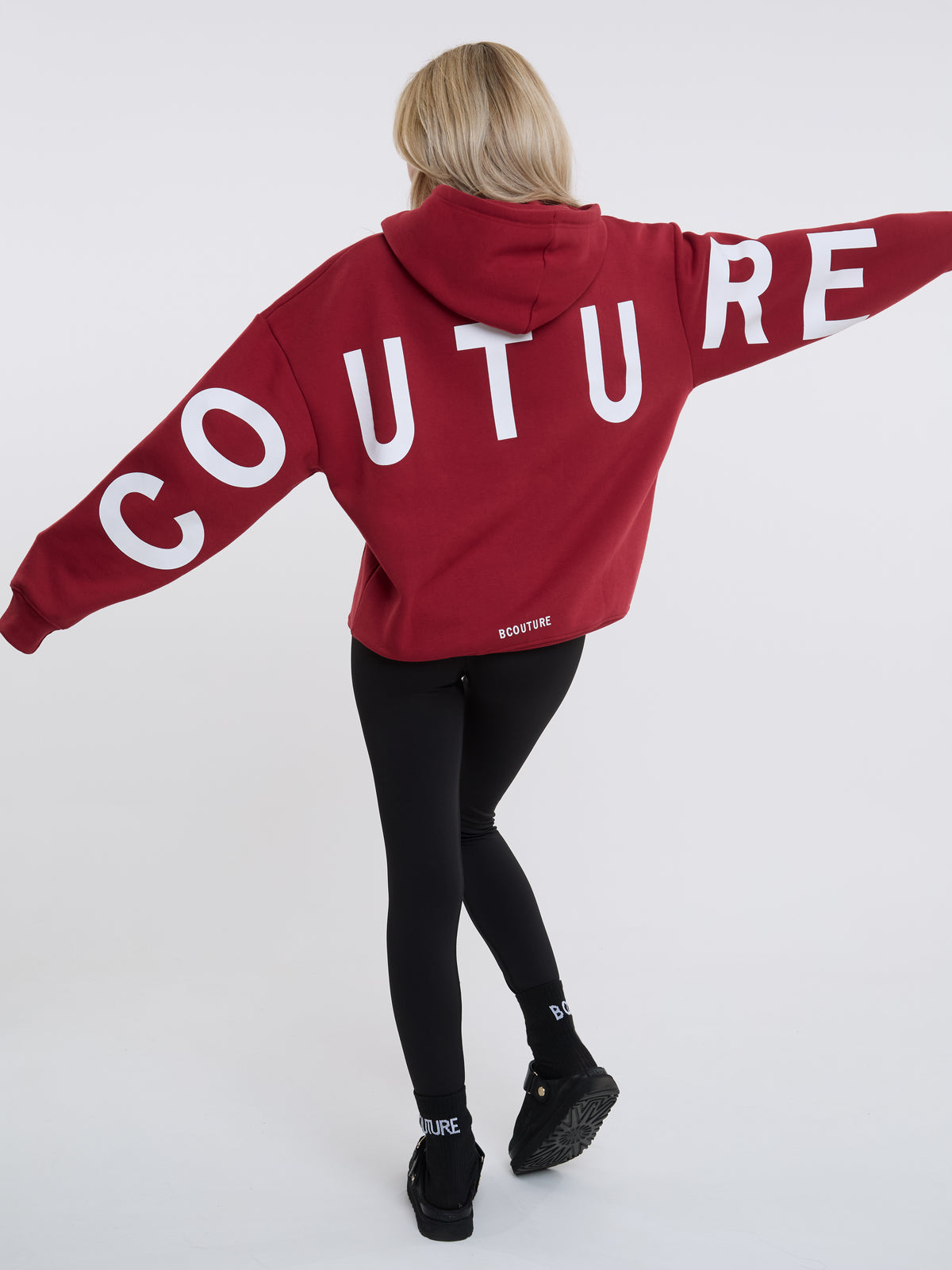 A woman viewed from behind wearing a cropped red hoodie with the word 'COUTURE' printed across the back in large white letters, paired with black leggings and black shoes, against a light gray studio background.