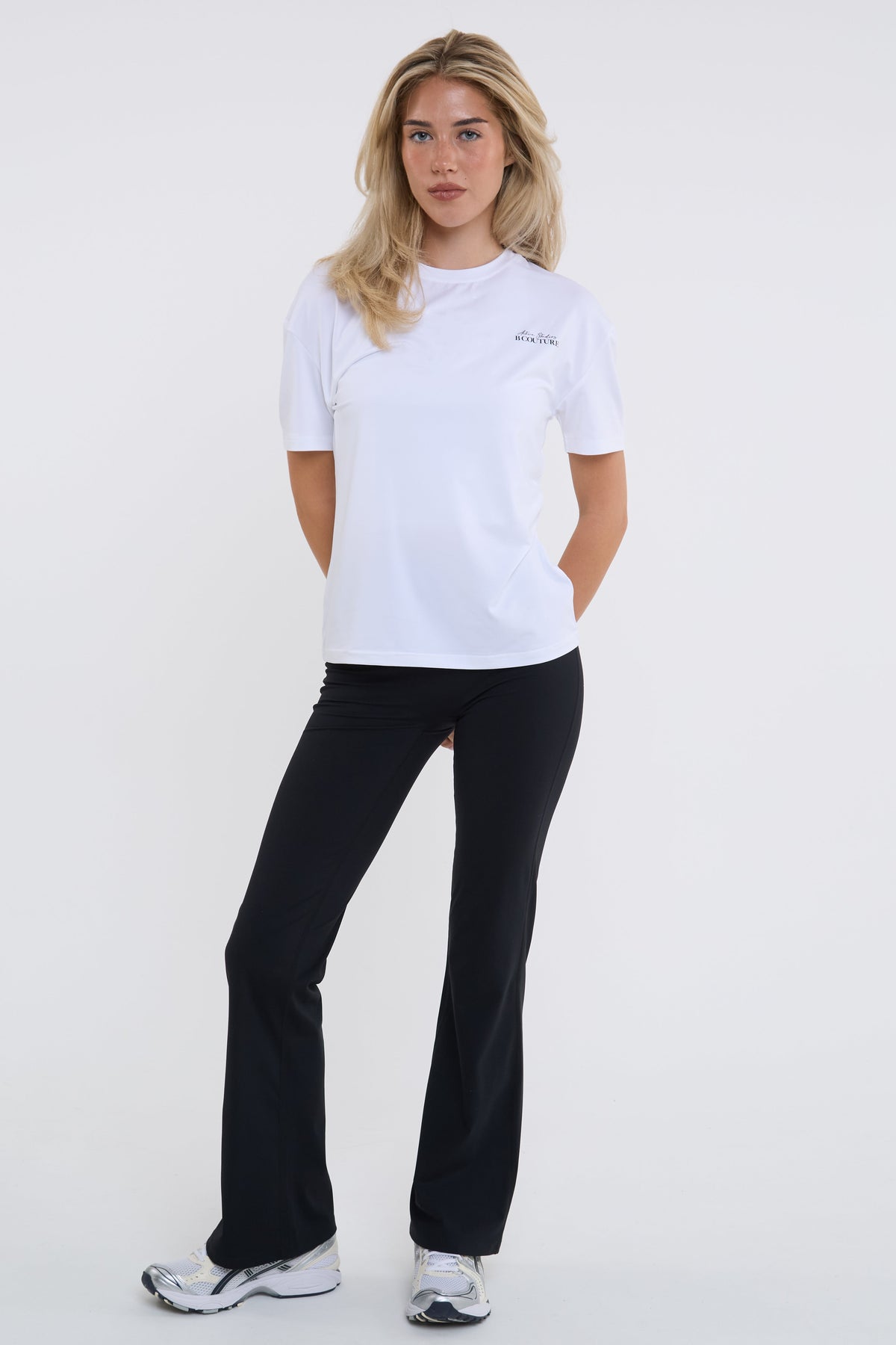 A woman modeling a white short-sleeve athletic t-shirt with a small script logo on the left chest, paired with black flared pants and white sneakers, against a plain white studio background.