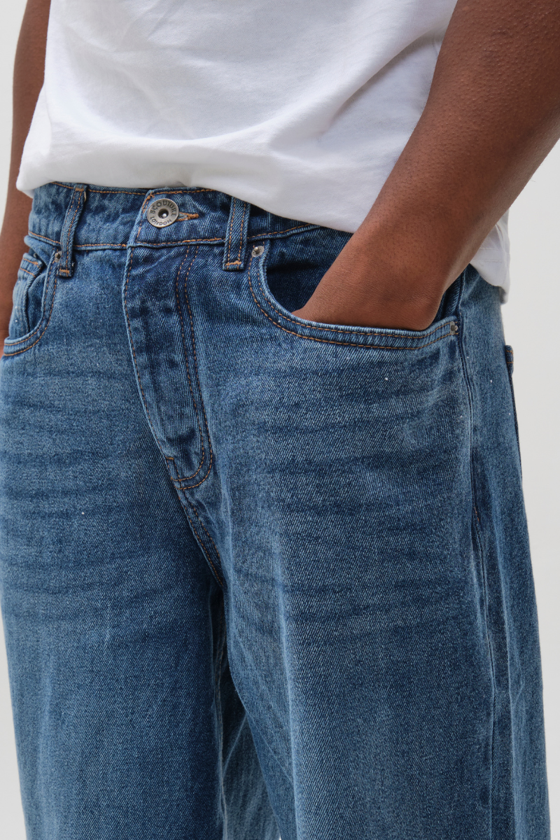 A person wears B Couture's Southbank Loose Fit Rigid Denim Jeans in Antique Blue, styled with a white t-shirt and both hands in their front pockets. The image highlights the waist and upper legs, emphasizing modern casual style.