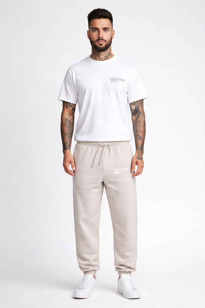 A man with tattoos and a groomed beard stands facing forward against a white background, wearing a white T-shirt, Heavy Weight Oversized Couture Department Joggers in grey with a drawstring, and white sneakers.