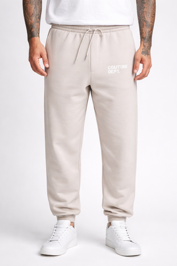 A person is wearing Heavy Weight Oversized Couture Department Joggers in grey, featuring "COUTURE DEPT." on the left thigh, styled with a white t-shirt and sneakers. Their tattooed arms are visible; the photo shows them from shoulders down.