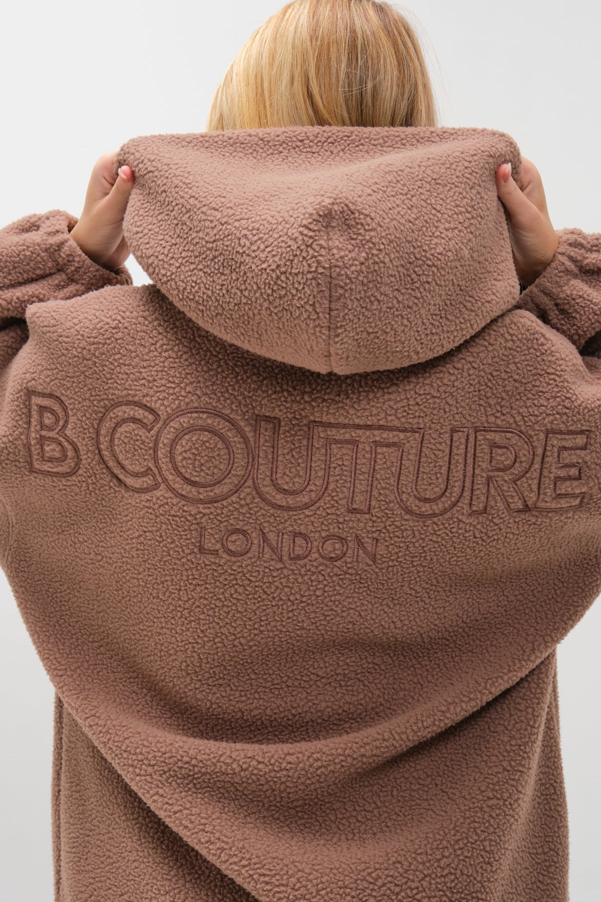 A person with blonde hair wears the Isla Sherpa Hoodie & Leggings Set in brown/beige, featuring a plush fleece, fuzzy hooded coat with "B COUTURE LONDON" embroidered on the back, holding the hood up for cozy warmth.