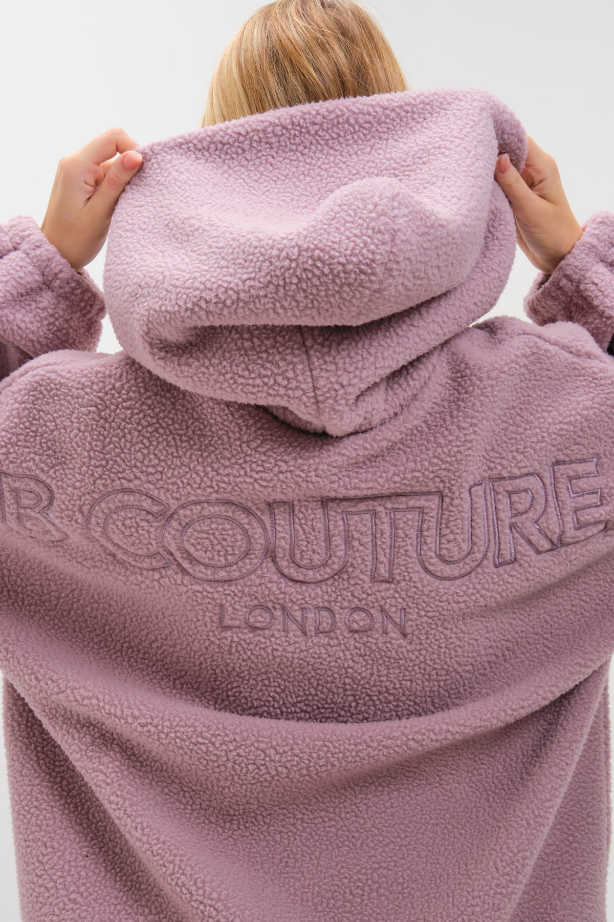 A person lifts the hood of the Isla Sherpa Hoodie in mauve, showing its cosy, textured fleece. Large "COUTURE LONDON" embroidery stands out on the back, while only their hands and hair are visible, highlighting this warm statement set.