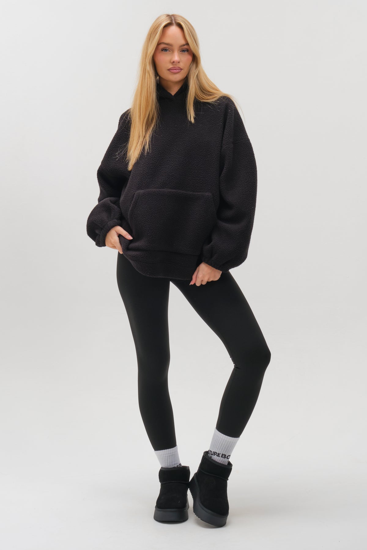 A woman with long blonde hair stands confidently against a white background, wearing the Isla Sherpa Hoodie & Leggings Set in black—an outfit that radiates cosy warmth and effortless style.