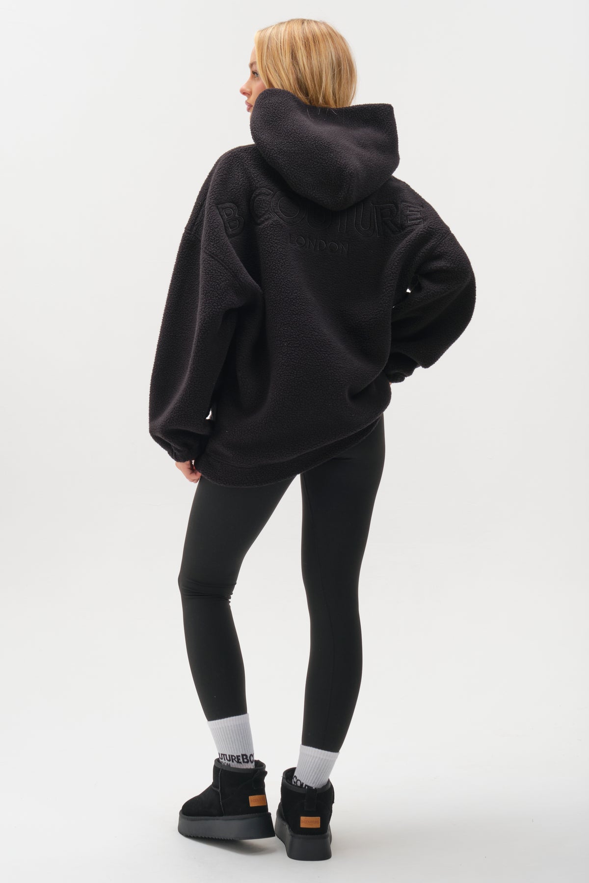 A person with long blonde hair faces away in the Isla Sherpa Hoodie & Leggings Set - Black, styled with white socks and black platform sneakers against a plain white background.
