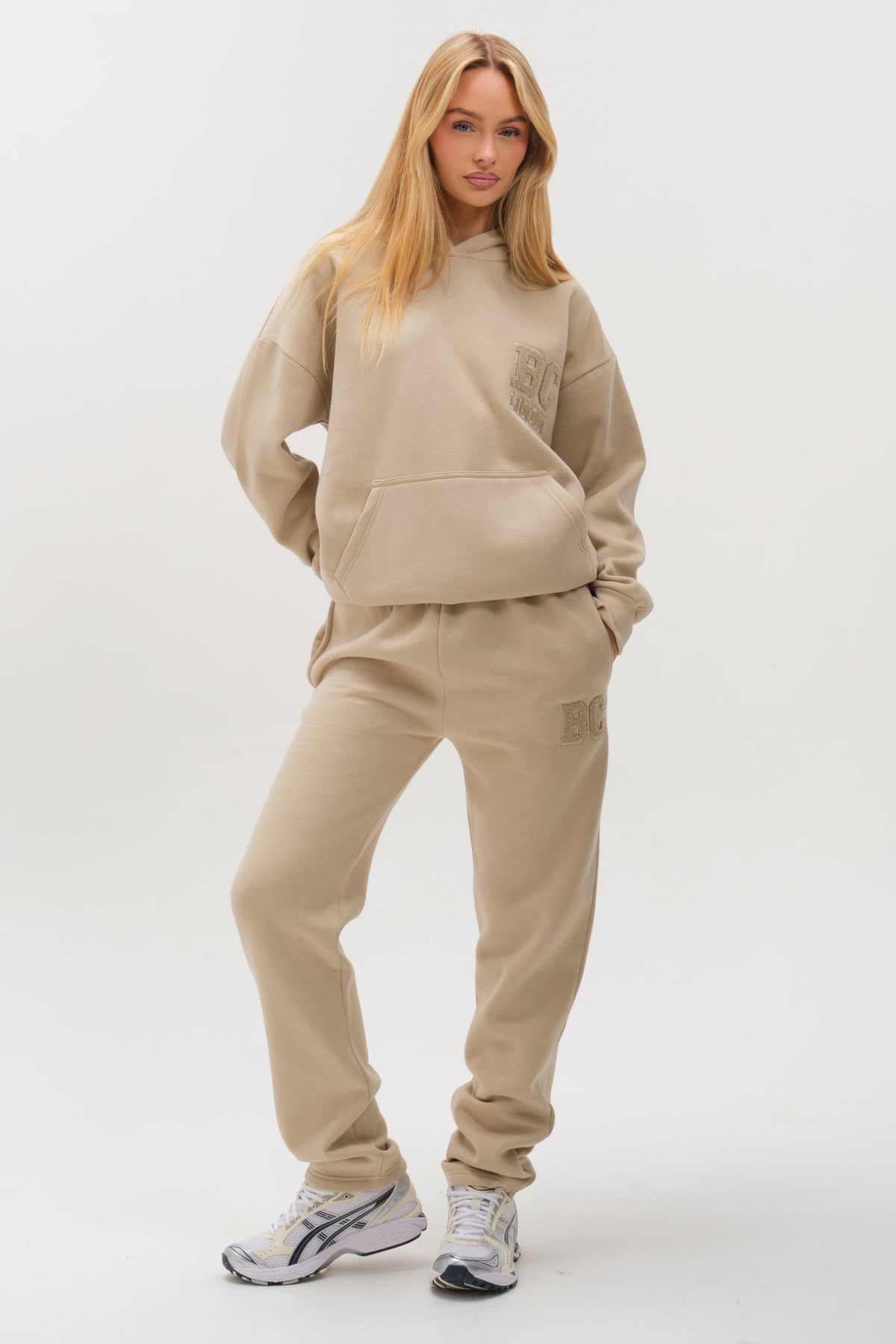 A woman with long blonde hair wears the Etty Fleece Tracksuit in Stone, styled in a relaxed fit with white sneakers. She stands with her hands in her pockets against a plain white background.