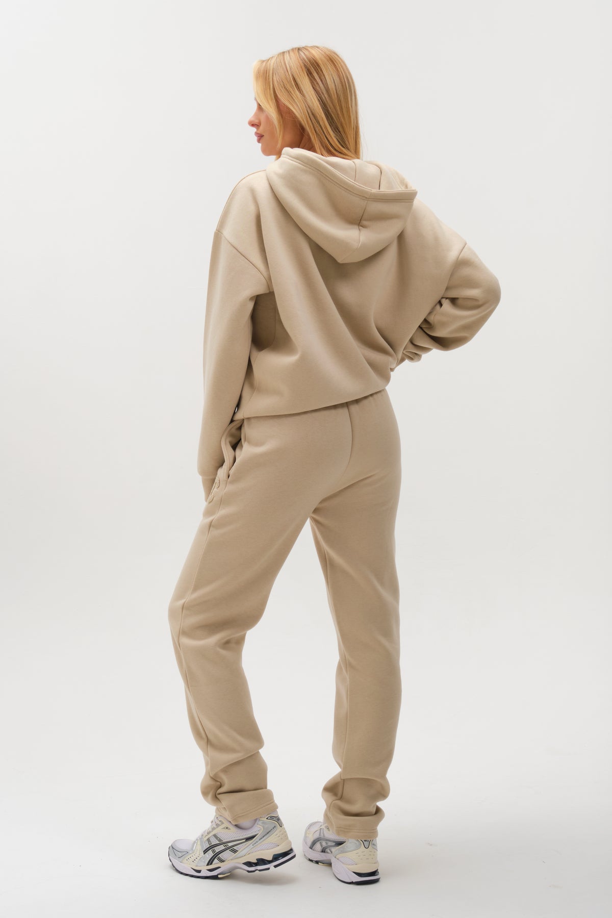 A person stands facing away, wearing the Etty Fleece Tracksuit in Stone—a relaxed fit beige hoodie and jogger set with white sneakers—set against a plain white background.