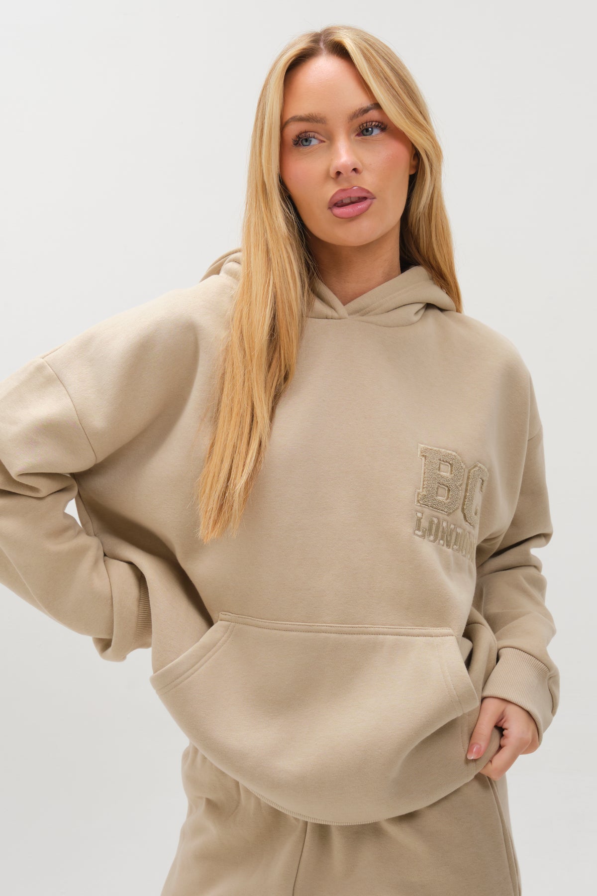A woman with long blonde hair poses against a light background in the Etty Fleece Tracksuit - Stone, a relaxed fit beige jogger set featuring a hooded sweatshirt with BC LONDON embroidery and matching pants.