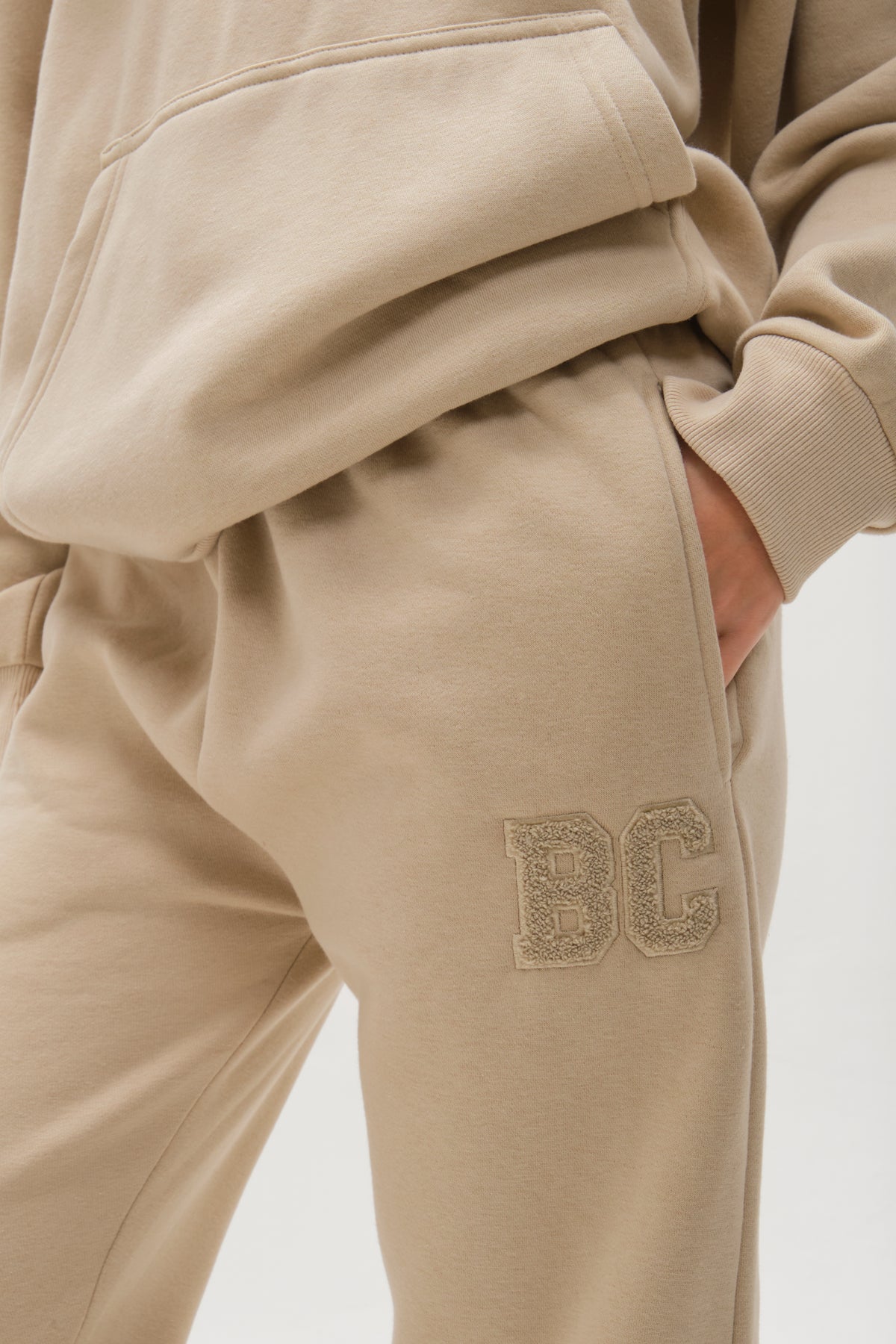 A person wears the Etty Fleece Tracksuit in Stone, a relaxed fit jogger set. One hand is in their pocket, and the sweatpants have textured "BC" embroidery near the hip.