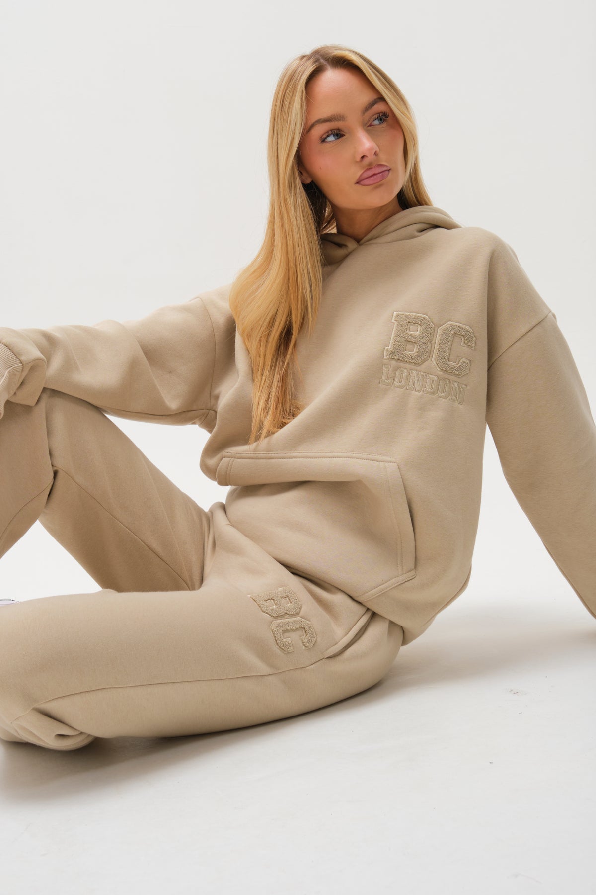 A woman with long blonde hair sits on the floor against a white background, wearing the Etty Fleece Tracksuit - Stone, a relaxed fit beige hoodie and matching joggers featuring BC London embroidery on the chest and leg.