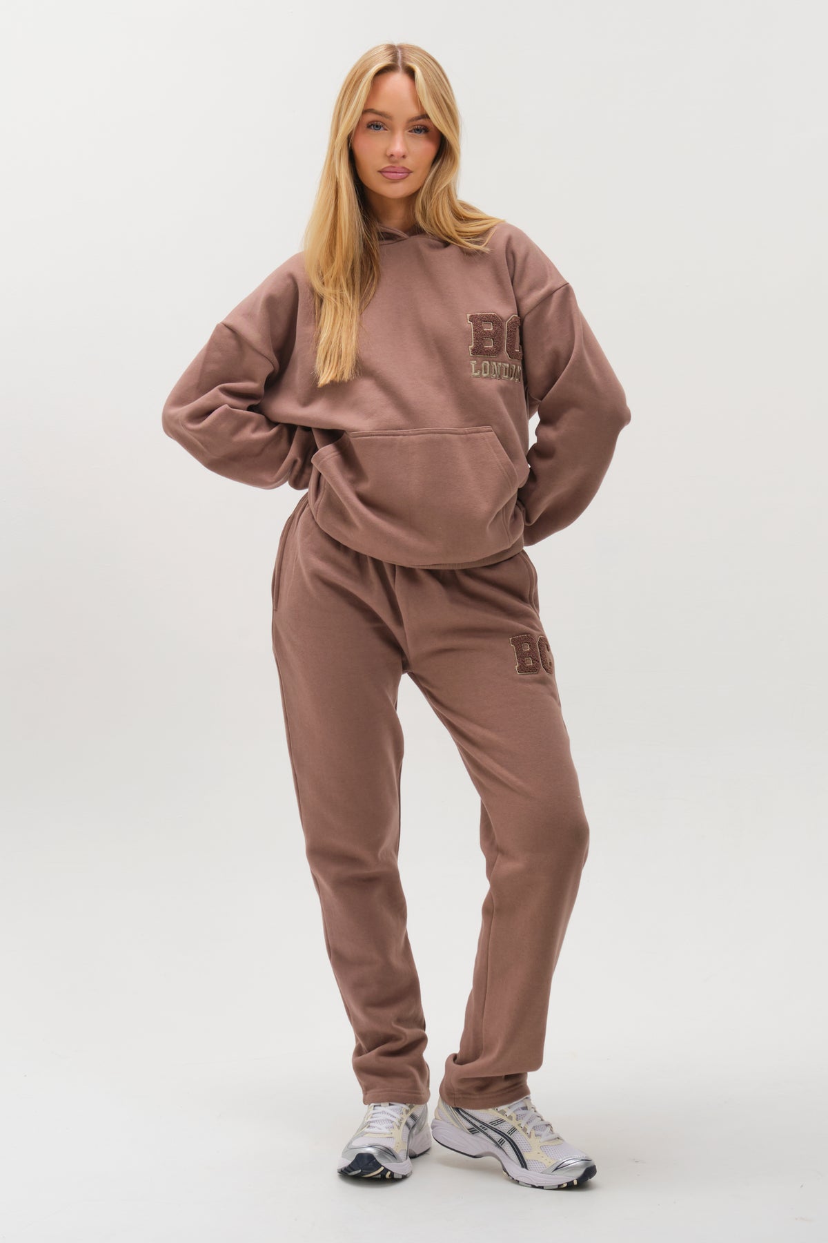 A woman with long blonde hair wears the Etty Fleece Tracksuit in Mink Brown, featuring a relaxed fit and BC London branding. She poses with hands in pockets and white sneakers against a plain white background.