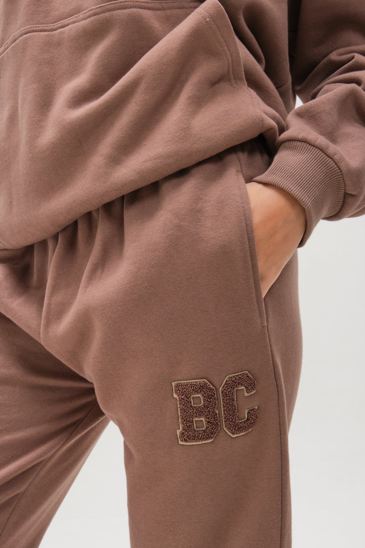 Someone wears the Etty Fleece Tracksuit in Mink Brown, with a relaxed fit and one hand in their pocket. The visible joggers feature textured BC lettering embroidered near the pocket; only the lower half of the outfit is shown.
