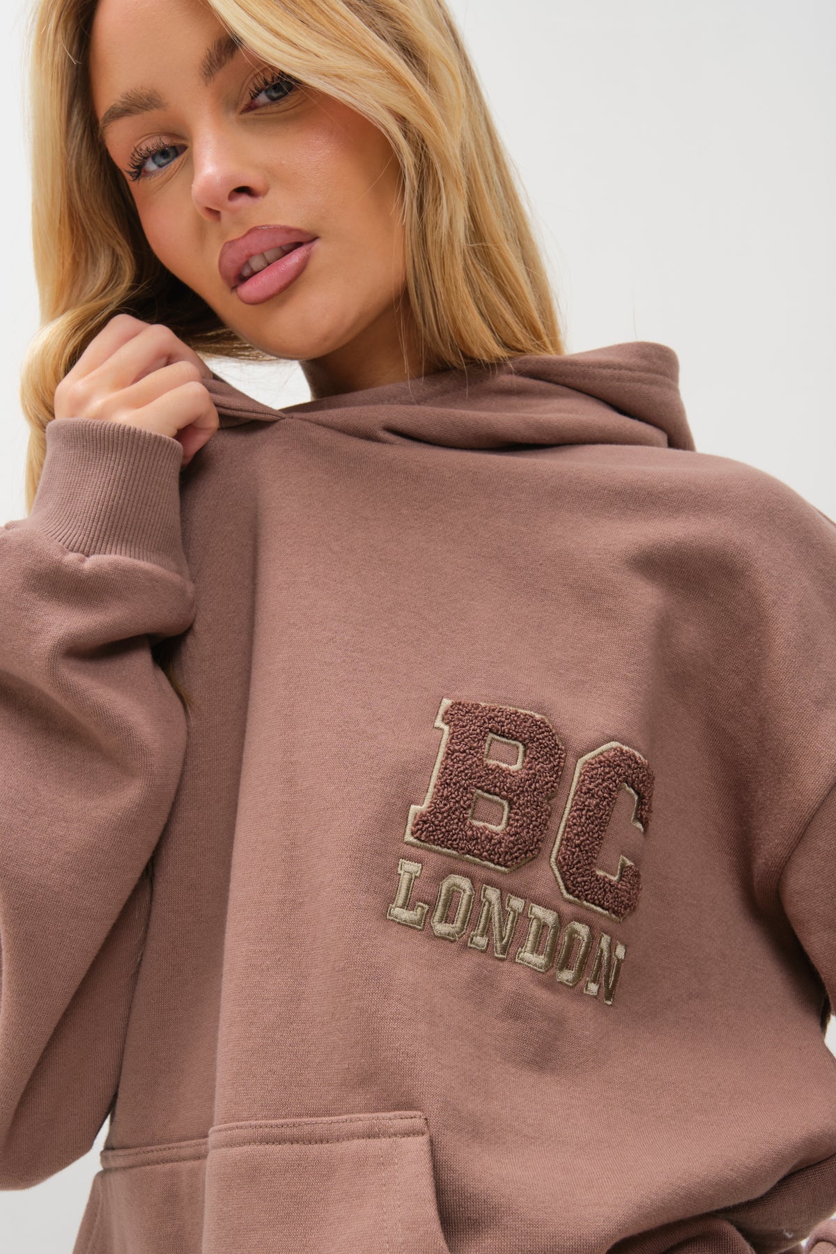 A woman with long blonde hair wears the Etty Fleece Tracksuit in mink brown, featuring BC LONDON embroidery on the front. She gently holds the collar and gazes at the camera against a plain light background.