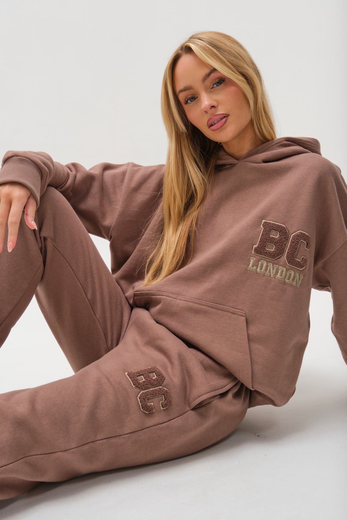 A woman with long blonde hair wears the Etty Fleece Tracksuit in Mink Brown, a relaxed fit hoodie and matching sweatpants with BC London printed on them, sitting on the floor against a plain white background.