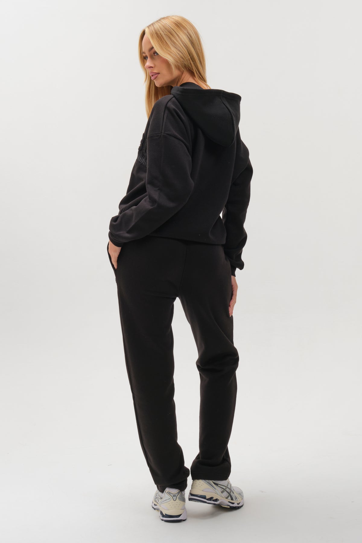 A woman with long blonde hair models the Etty Fleece Tracksuit - Black, featuring a relaxed fit hoodie and joggers, hands in pockets, wearing white sneakers against a plain white background.