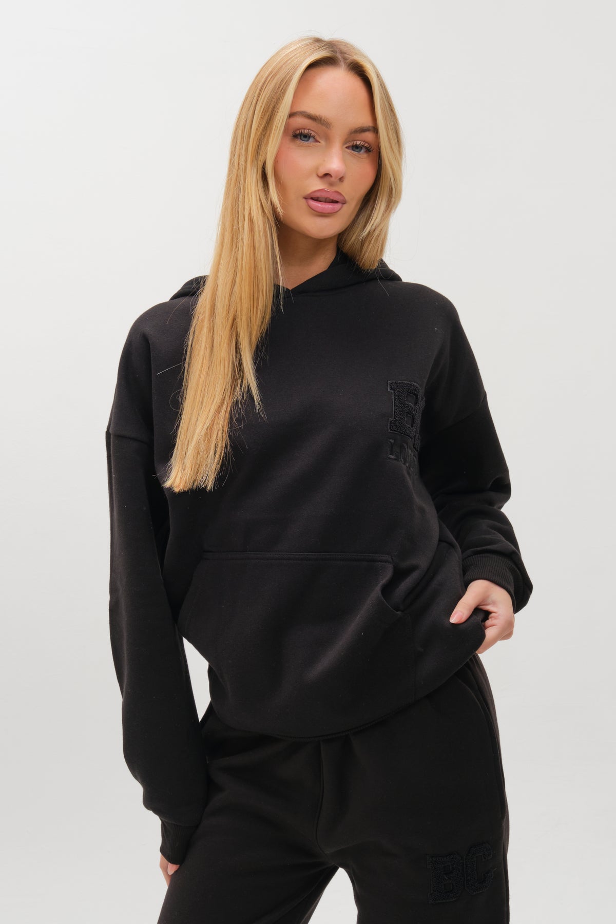 A woman with long blonde hair models the Etty Fleece Tracksuit in black, featuring a relaxed fit hoodie and matching joggers, standing with one hand in her pocket against a plain light background.