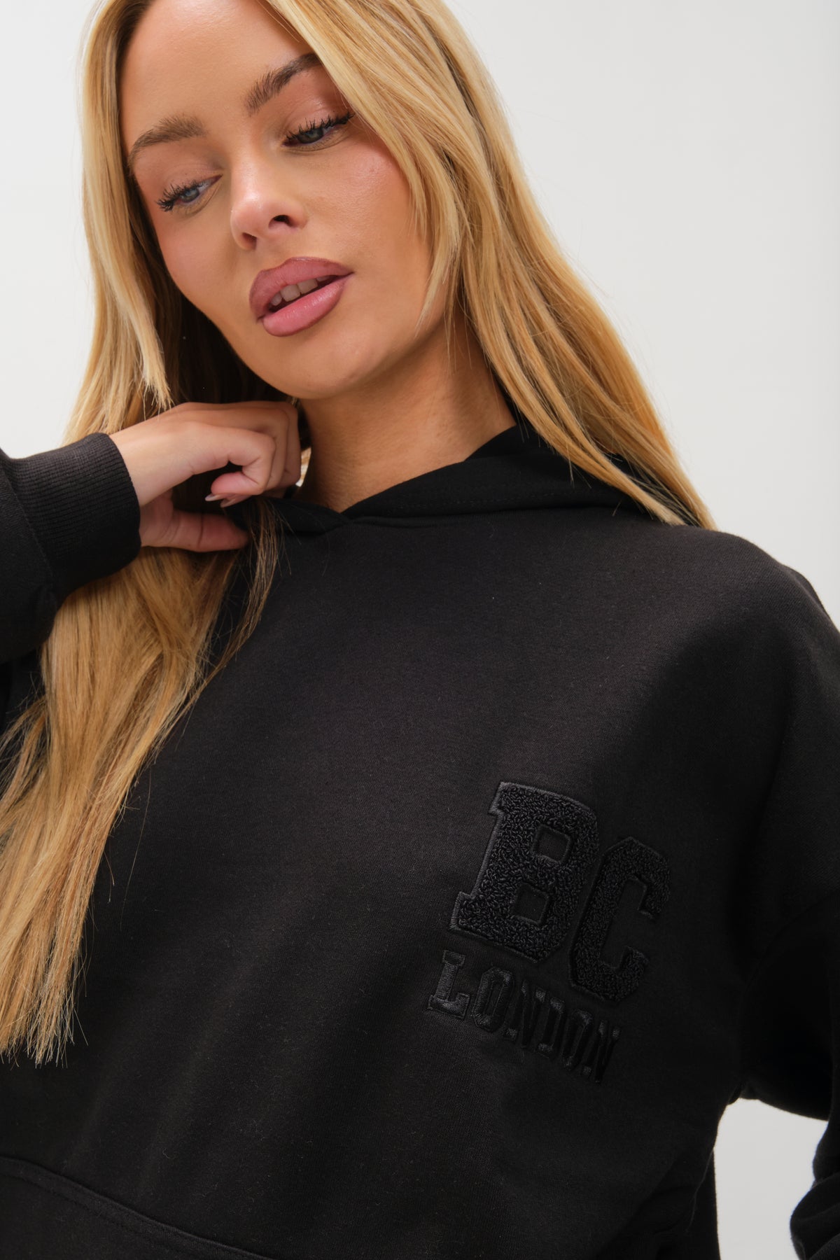 A woman with long blonde hair wears the Etty Fleece Tracksuit - Black, featuring a relaxed fit and BC LONDON embroidery on the chest. She looks downwards, her hand gently touching her neck.