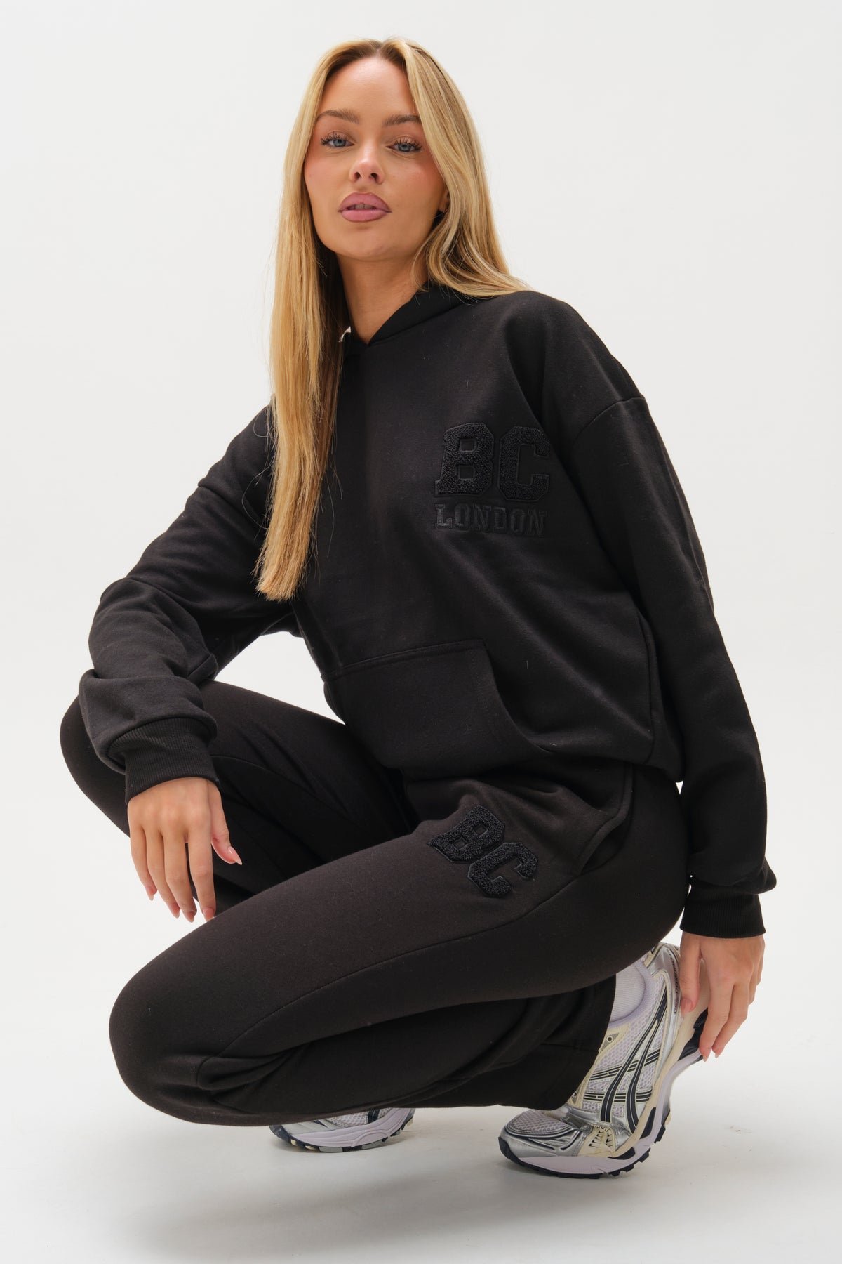 A woman with long blonde hair wears the Etty Fleece Tracksuit in black, featuring a relaxed fit hoodie and matching joggers, paired with silver and white athletic sneakers against a plain white background.