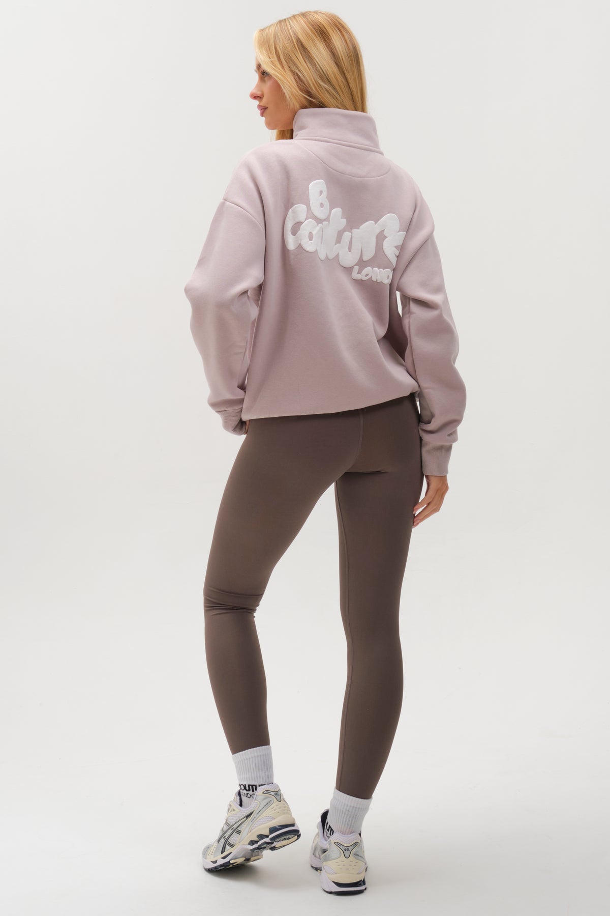 A woman faces away, wearing the Hatherley Half-Zip Sweatshirt & Leggings Set in Stone, paired with white socks and sneakers for a cosy look against a plain white background.