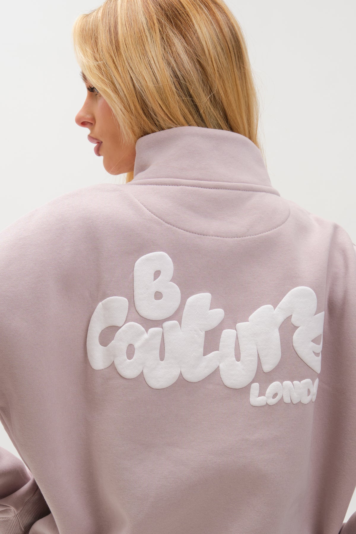 A woman with blonde hair models the Hatherley Half-Zip Sweatshirt & Leggings Set in Stone, turned slightly away to show the relaxed fit and large white "B Couture LONDON" text printed on the back.