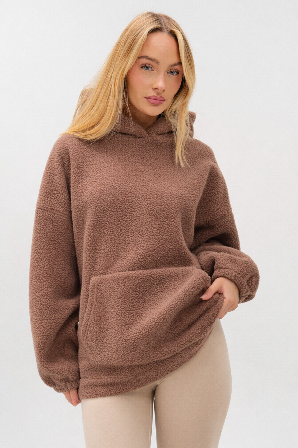 A woman with long blonde hair models the Isla Sherpa Hoodie & Leggings Set in brown and beige, standing against a plain light background with one hand in her pocket, exuding cozy warmth.