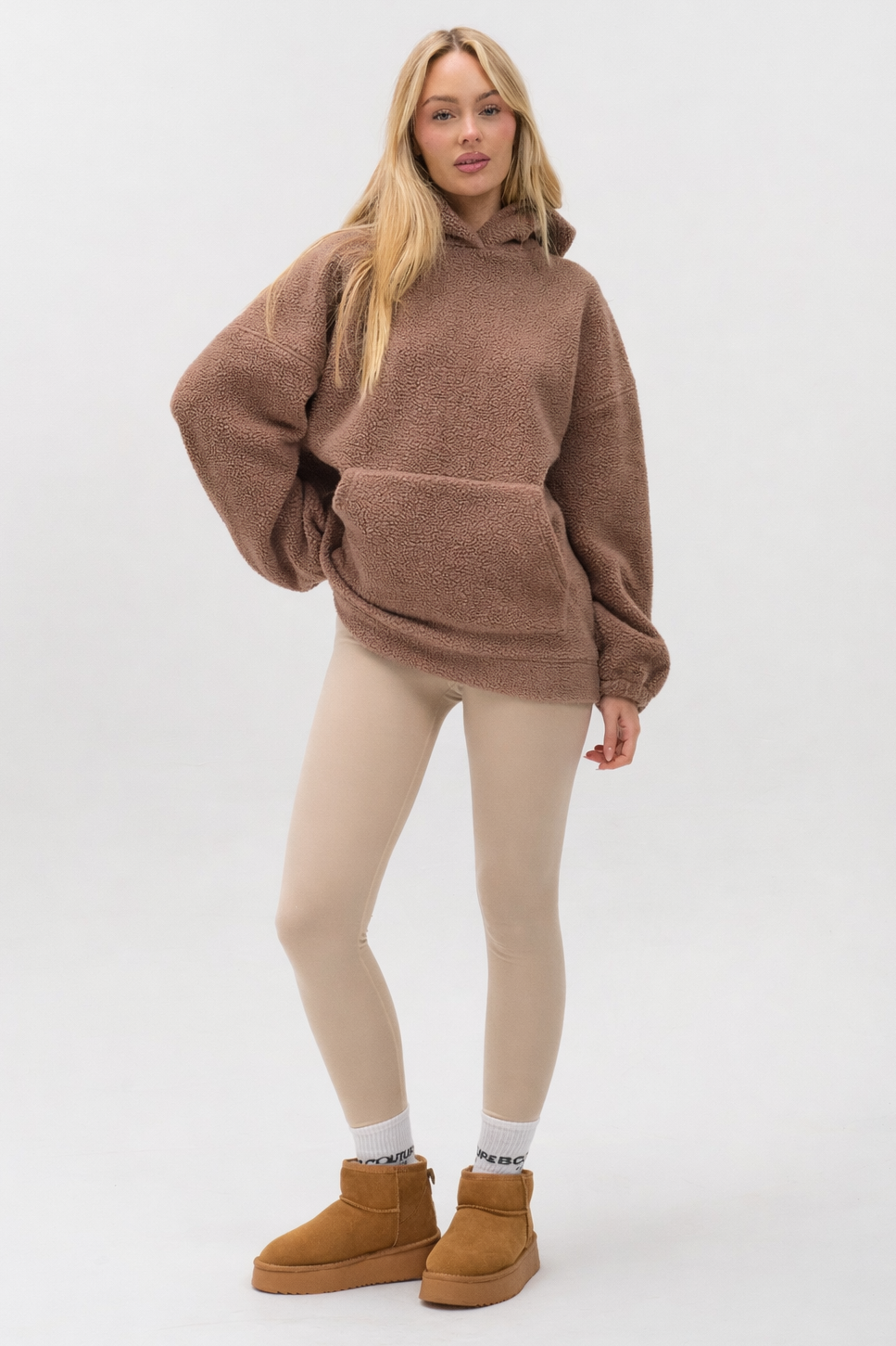 A woman with long blonde hair stands in a relaxed pose wearing the Isla Sherpa Hoodie & Leggings Set in brown/beige, paired with gray socks and tan ankle boots, radiating cozy warmth against a plain background.