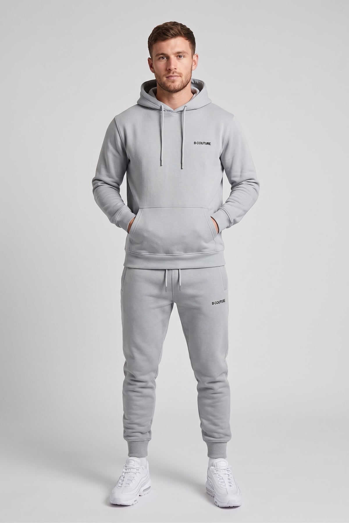 A man modeling a light gray two-piece tracksuit consisting of a pullover hoodie and matching jogger pants, with white sneakers, against a solid light gray studio background.