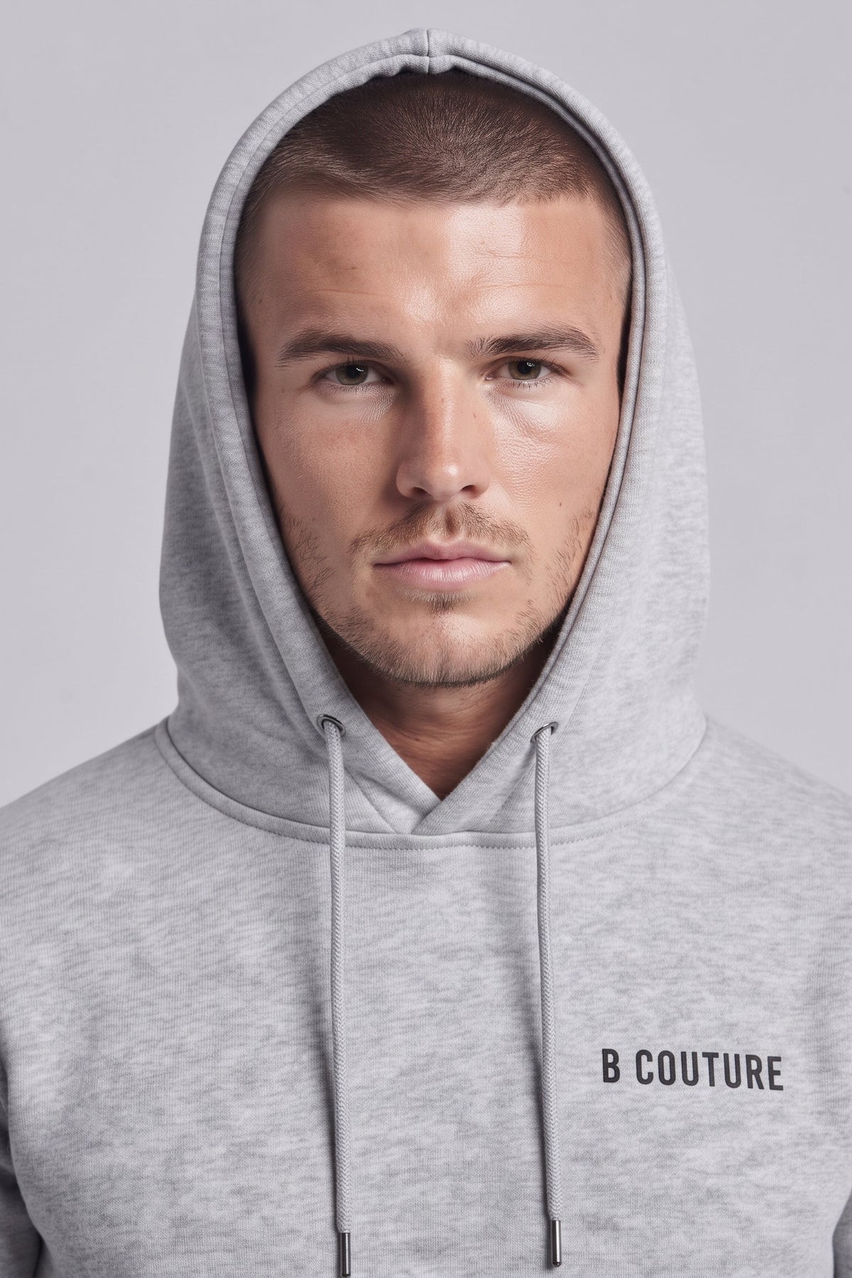 A man wearing a light gray heather pullover hoodie with 'B COUTURE' printed in black on the left chest, shown against a solid light gray background.