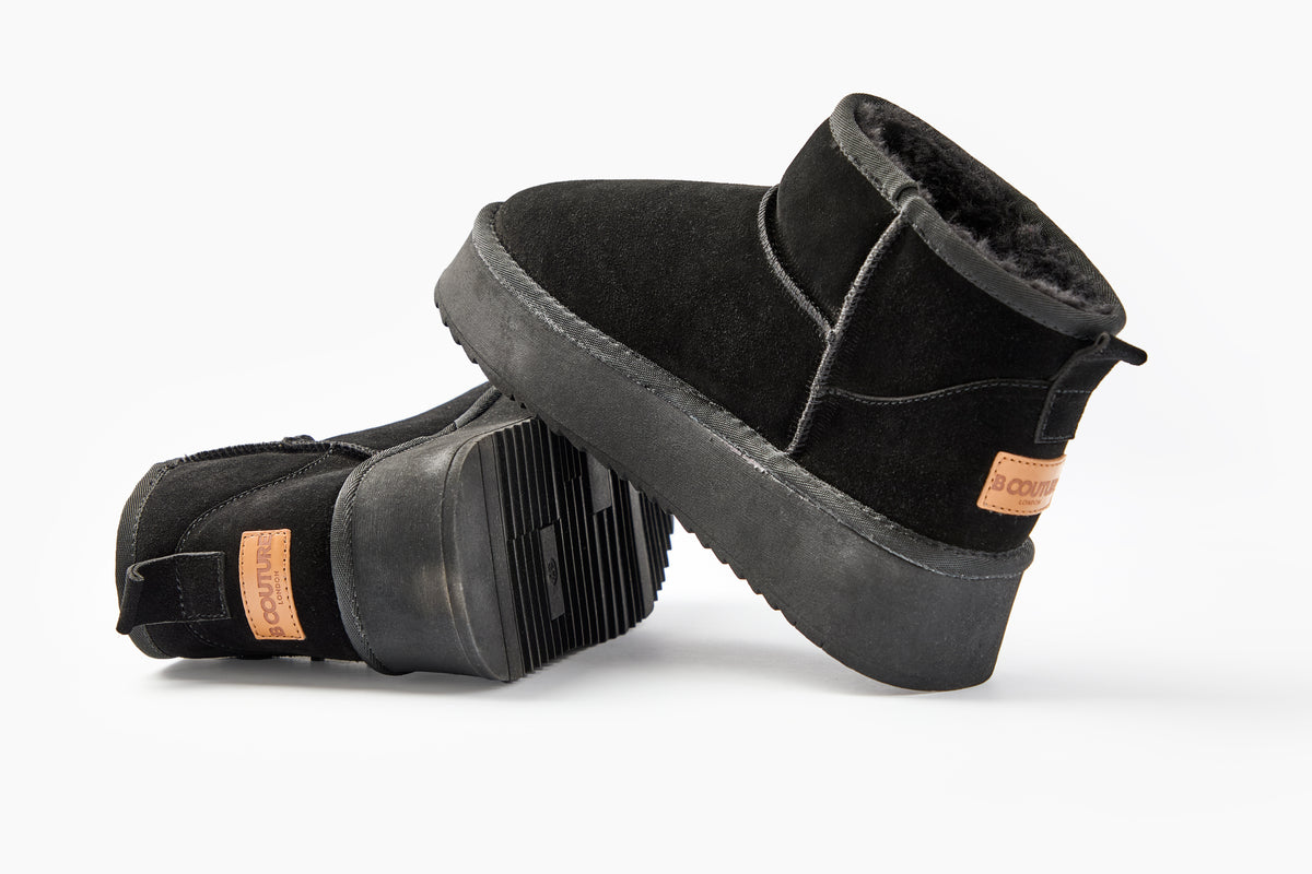 A pair of Amwell Genuine Suede Mini Boots in black, featuring platform rubber soles and tan leather logo patches on the back, is displayed against a white background. One boot stands upright while the other leans to reveal its sole.