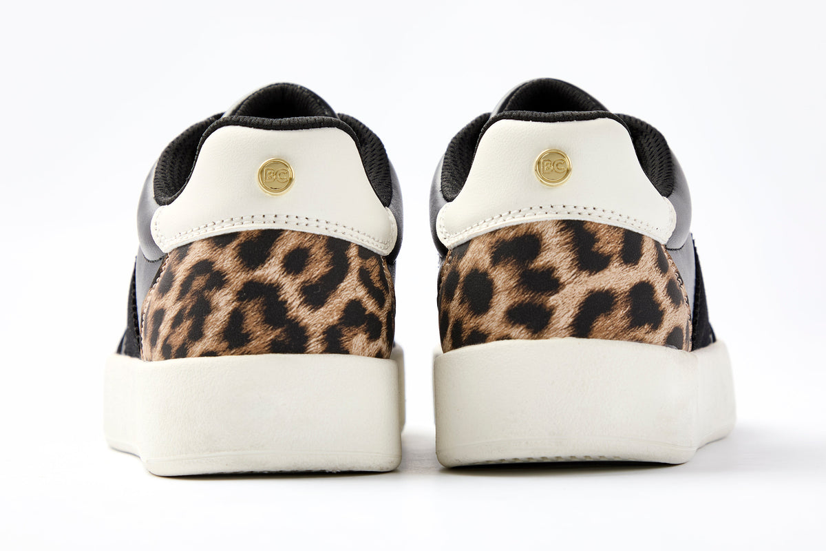The Ampton Low-Profile Trainer in Black/Leopard features black and white uppers, white soles, brown leopard-print accents on the heels, and a small gold circular logo on each heel tab.
