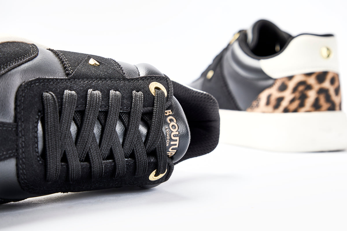 Close-up of the Ampton Low-Profile Trainer in black leather with gold eyelets, branded tongue, white sole, and leopard print heel detail, set against a white background.