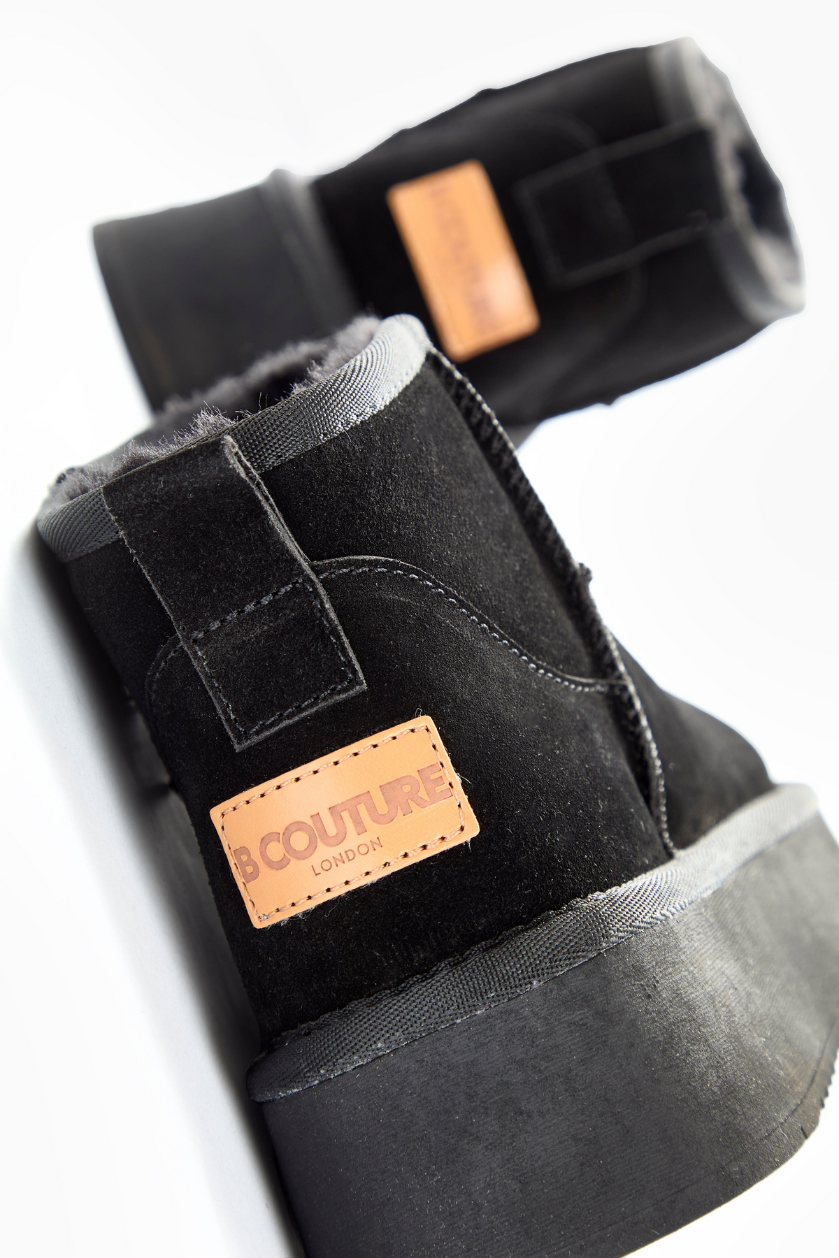 A close-up of Amwell Genuine Suede Mini Boots in black with thick platform rubber soles and a tan leather COUTURE LONDON patch stitched on the side, shown diagonally on a white background.