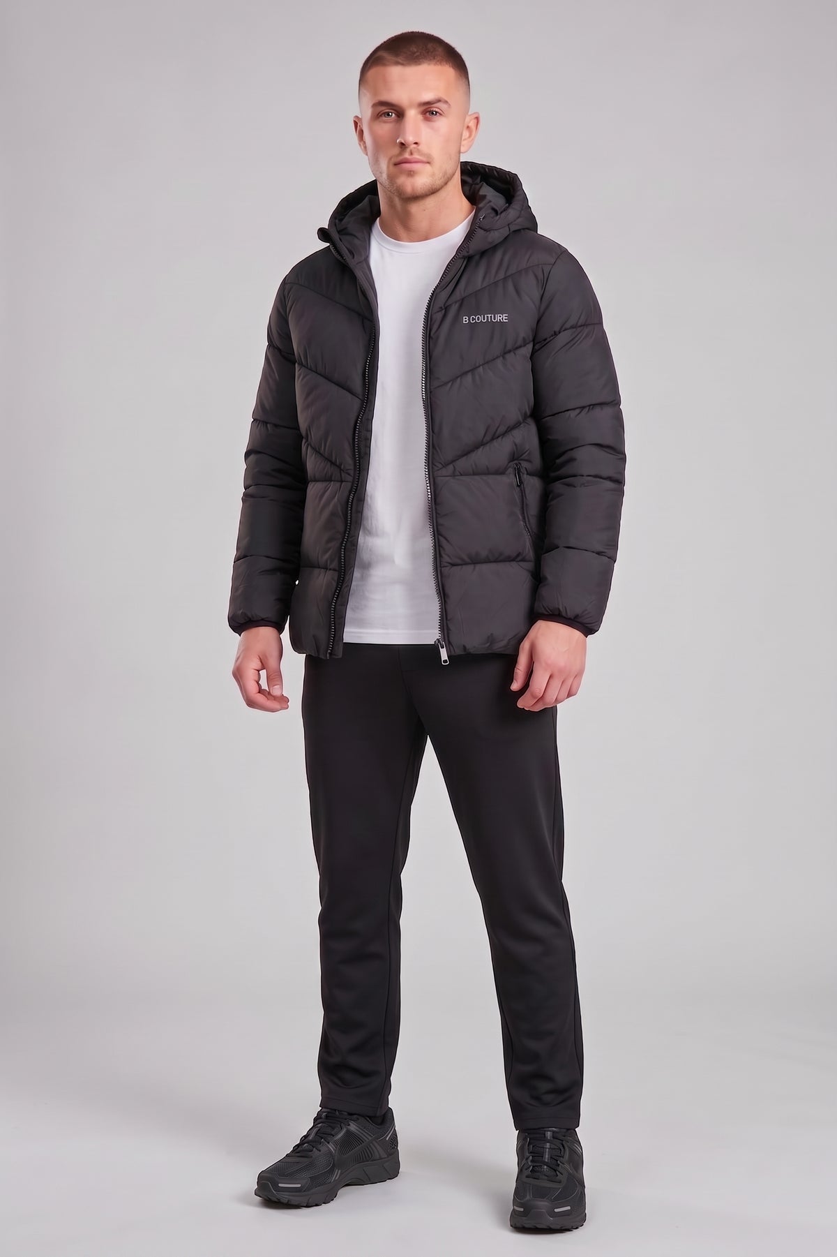 A man modeling a black hooded puffer jacket over a white t-shirt, black pants, and black shoes, standing against a light gray studio background.